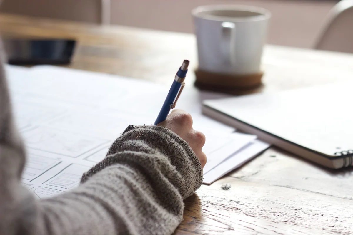 person writing on a book on a wooden table with ceramic mug